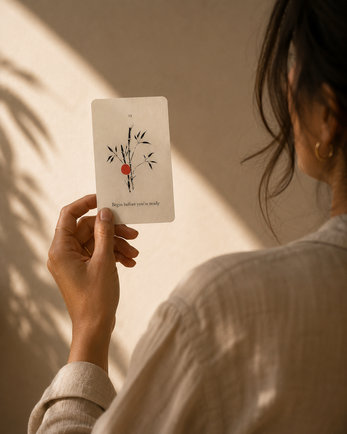 A person in a beige linen shirt holds up the Kokū Begin before you're ready card, a hand-painted bamboo illustration with a vermilion dot, against a warm beige wall with soft leaf shadows.