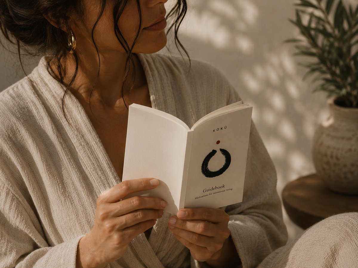 A woman in a beige robe holds the open Kokū Pocket Book in soft indoor light, with a potted plant behind her.